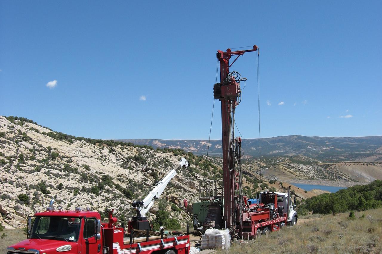 A truck-mounted drilling rig raising its mast on a sagebrush ridge above a reservoir.